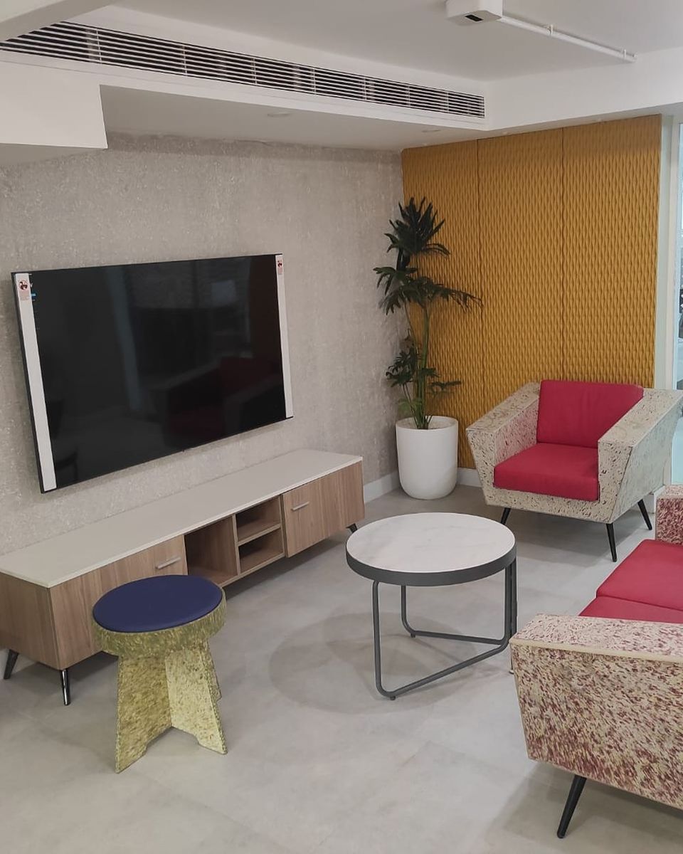 An office lounge with a wall-mounted screen, red-upholstered seating and a yellow textured wall panel.